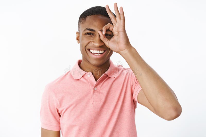 Portrait of bright and cheerful young dark-skinned guy in pink polo shirt showing okay or zero gesture over eye looking stock image Portrait of bright and cheerful young dark-skinned guy in pink polo shirt showing okay or zero gesture over eye looking stock image