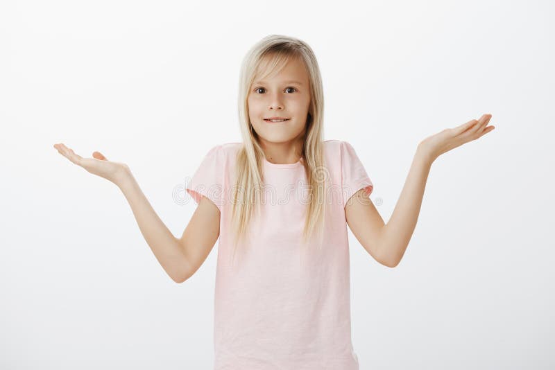 Portrait of charming female child with natural blond hair, smiling awkwardly and shrugging with spread palms, being questioned and unaware what is happening, having no idea how to answer on question.