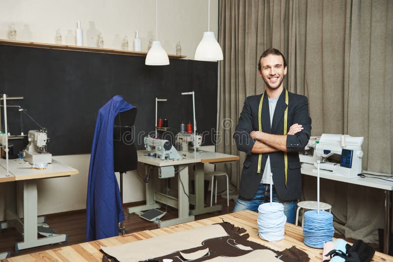 Portrait of cheerful good-looking male clothes designer with dark hair in fashionable outfit standing in workshop.