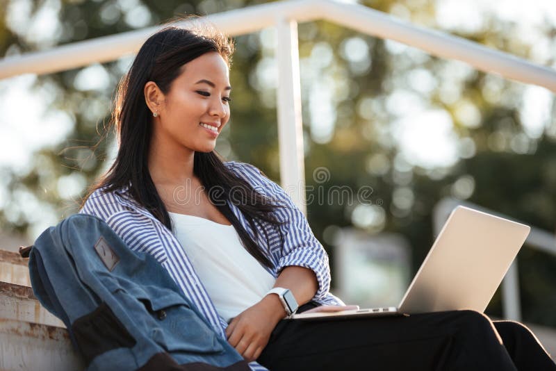 Portrait of a cheerful pretty asian female student holding laptop computer on her lap while sitting on the bench at the park