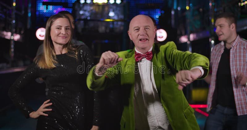 Portrait of cheerful senior Caucasian man and young pretty woman dancing in night club and looking at camera. Joyful old