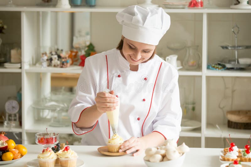 Portrait of smiling female professional confectioner topping a cupcake with cream using a pastry bag. Various delicious cakes lying on the table. Lifestyle. Portrait of smiling female professional confectioner topping a cupcake with cream using a pastry bag. Various delicious cakes lying on the table. Lifestyle