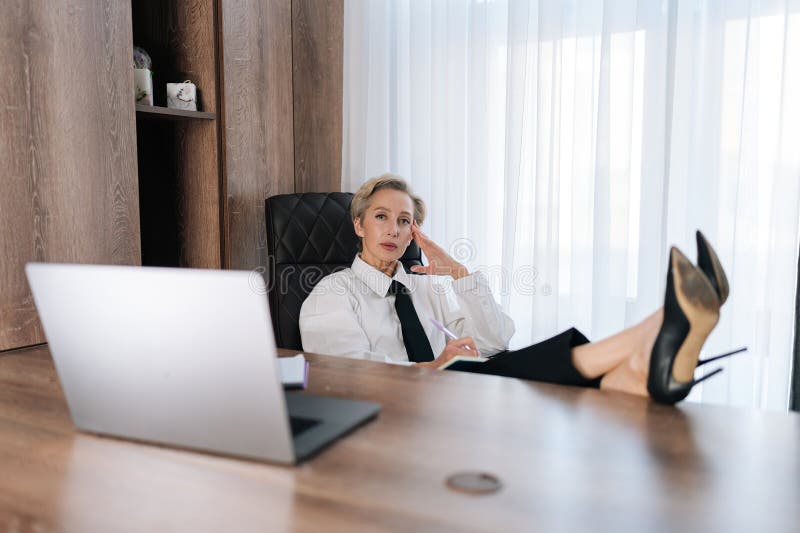 Portrait of confident middle-aged female CEO putting legs on desk with laptop, confident looking at camera. Successful business woman relaxing in cabinet. Concept relaxation at workplace