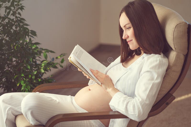 Portrait confortable franc d'une femme enceinte se reposant à la maison et lisant un livre sur une chaise de basculage par la fen images libres de droits Portrait confortable franc d'une femme enceinte se reposant à la maison et lisant un livre sur une chaise de basculage par la fen images libres de droits