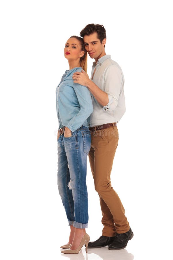 Portrait of couple embraced in studio background. she is looking at the camera with both hands in pockets while he is holding her from behind.