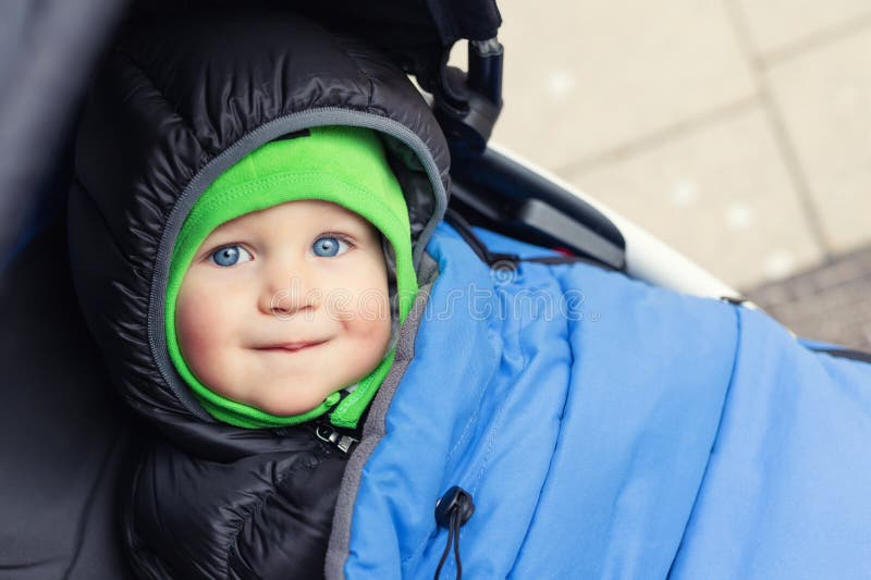 Portrait of cute caucasian toddler boy in black hooded warm insulated dawn overall jacket and blue blanket sitting in stroller and