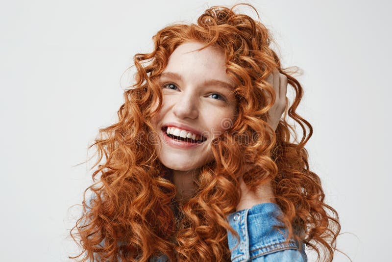 Portrait of cute happy girl smiling touching her curly red hair over white background.