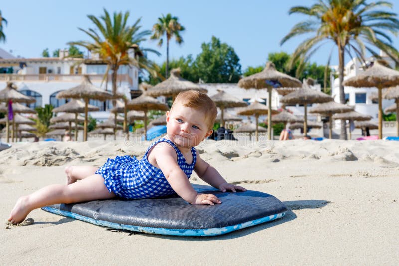 Portrait of cute little baby girl in swim suit on beach in summer. Adorable child having fun on surf board. Baby fashion for swimsuit.