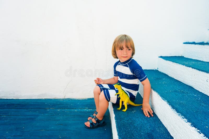 Adorable little boy playing with a dinosaur toy, wearing funny swim suit and beach shoes, sitting on blue stairs, summer vacations concept. Adorable little boy playing with a dinosaur toy, wearing funny swim suit and beach shoes, sitting on blue stairs, summer vacations concept