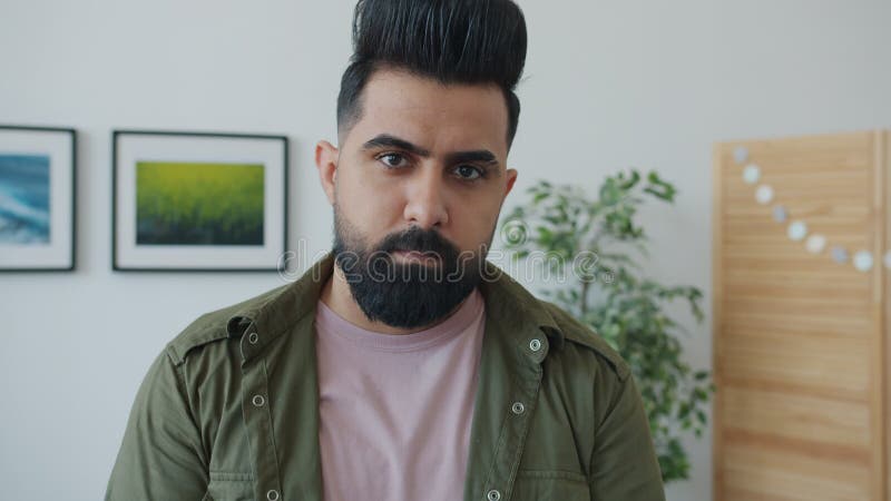 Portrait d'homme de race mixte barbu regardant la caméra avec le visage sérieux debout à la maison banque de vidéos