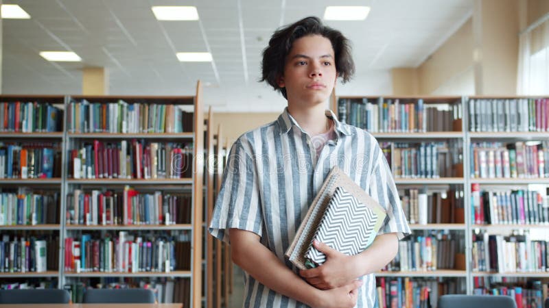 Portrait d'un adolescent sérieux marchant dans une bibliothèque scolaire avec des livres seuls banque de vidéos