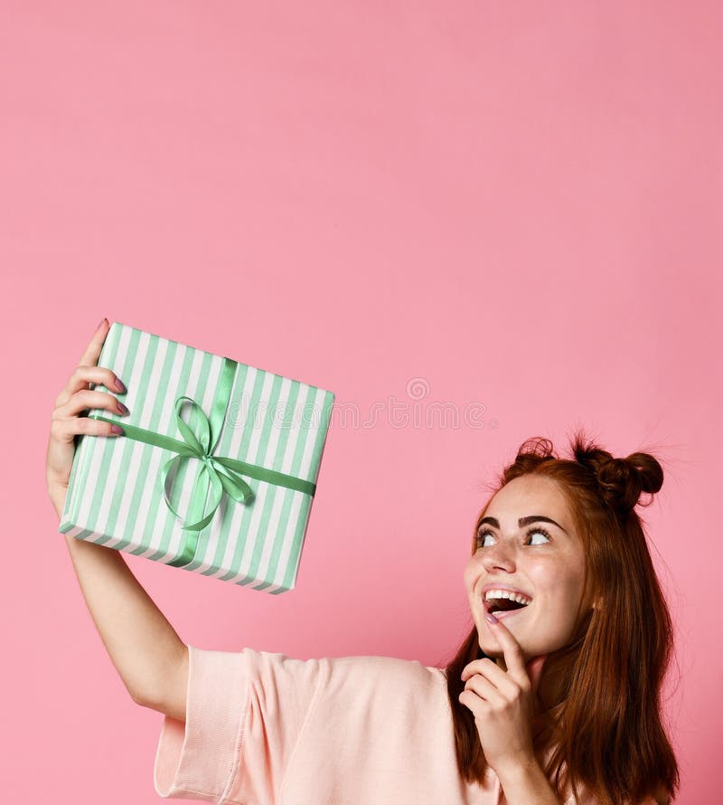 Portrait d'un boîte-cadeau roux assez de sourire et de le regarder de participation de fille, d'isolement au-dessus du fond rose image stock Portrait d'un boîte-cadeau roux assez de sourire et de le regarder de participation de fille, d'isolement au-dessus du fond rose image stock