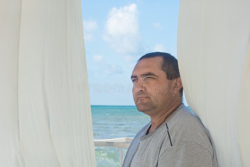 Portrait d'un homme de 40 ans à une fenêtre ouverte avec des rideaux blancs sur fond de mer ou d'océan regarde sur le côté images libres de droits