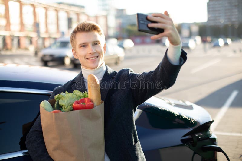 Portrait of young handsome european man, making selfie photo on his smartphone while leaning on his electric car, charging the battery at city power station after buying frsh products in trade mall. Portrait of young handsome european man, making selfie photo on his smartphone while leaning on his electric car, charging the battery at city power station after buying frsh products in trade mall.