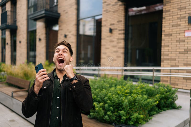Portrait d'un jeune homme heureux excité levant la main dans la victoire dehors tenant smartphone devant le moderne urbain images stock