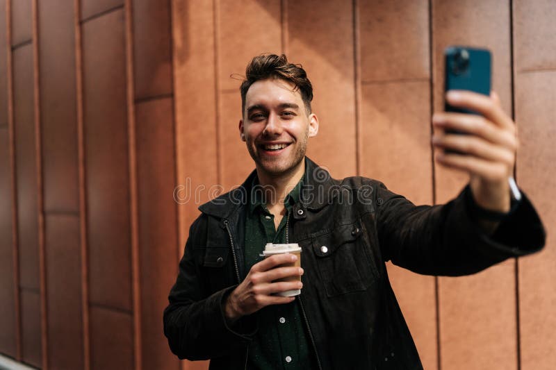 Portrait d'un jeune homme souriant et détendu qui prend un café tout en faisant un appel vidéo sur smartphone debout dans la rue u photos libres de droits