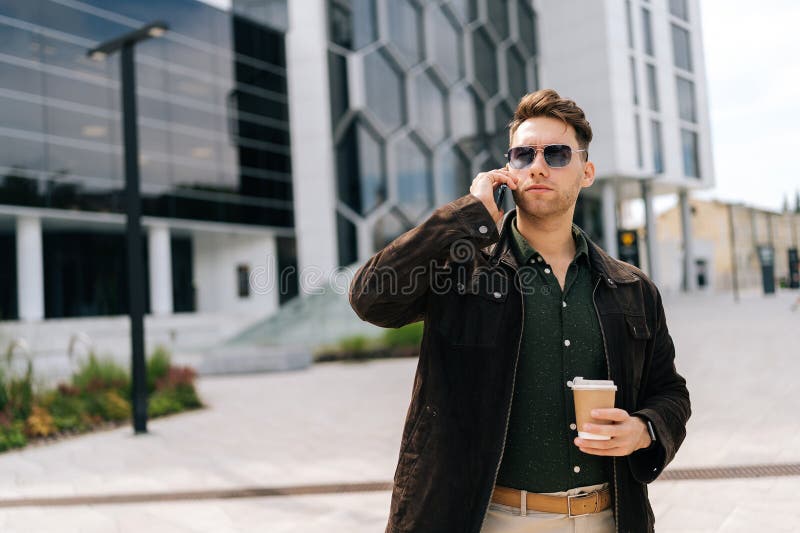 Portrait d'un jeune homme à barbe joyeux en tenue décontractée qui attend devant un bâtiment moderne qui parle sur smartphone et. Portrait de l'homme d'affaires confiant portant des lunettes de soleil marchant devant un immeuble de bureau moderne parlant sur son smartphone et tenant une tasse de café à emporter à l'air occupé et concentré