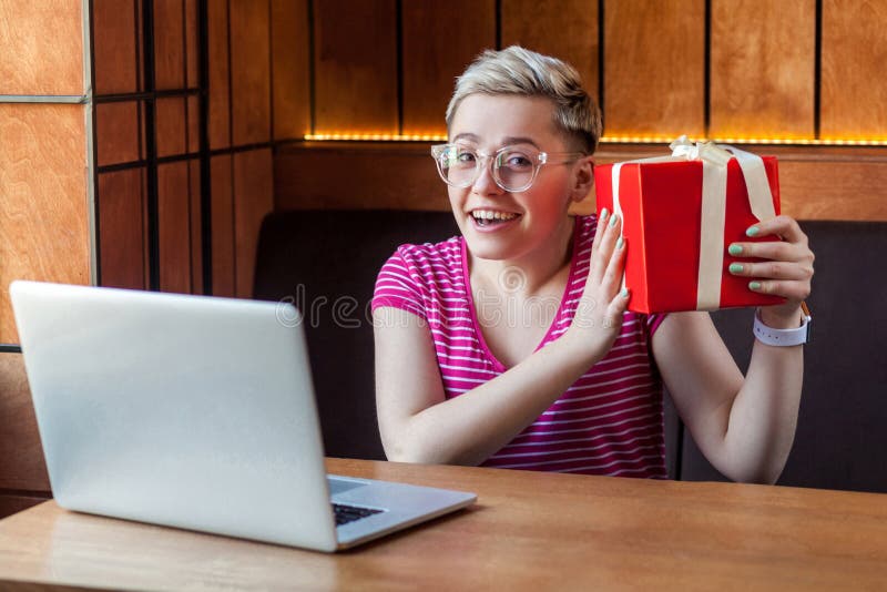 Portrait d'une belle jeune femme heureuse aux cheveux courts en t-shirt rose et lunettes est assise dans un café, tenant un cadea photo libre de droits
