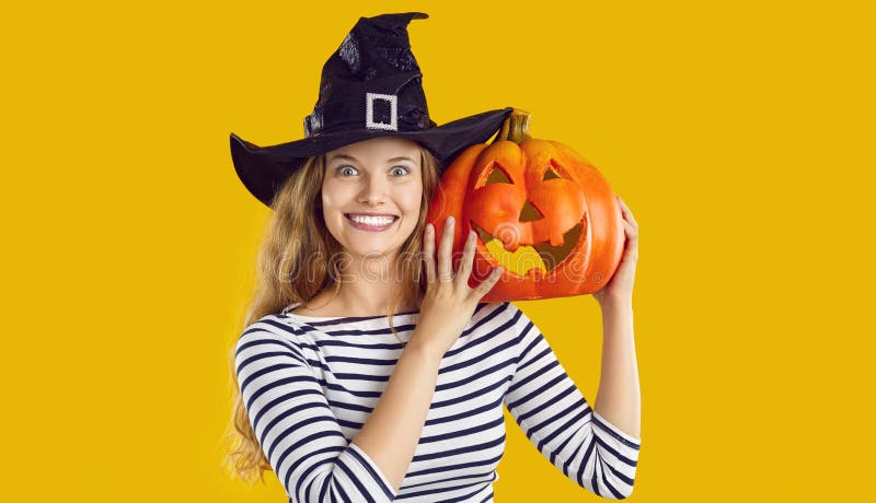 Portrait of a happy woman with a pumpkin. Beautiful young girl in a witch hat standing isolated on a yellow background, holding an orange pumpkin, looking at the camera and smiling. Halloween concept. Portrait of a happy woman with a pumpkin. Beautiful young girl in a witch hat standing isolated on a yellow background, holding an orange pumpkin, looking at the camera and smiling. Halloween concept