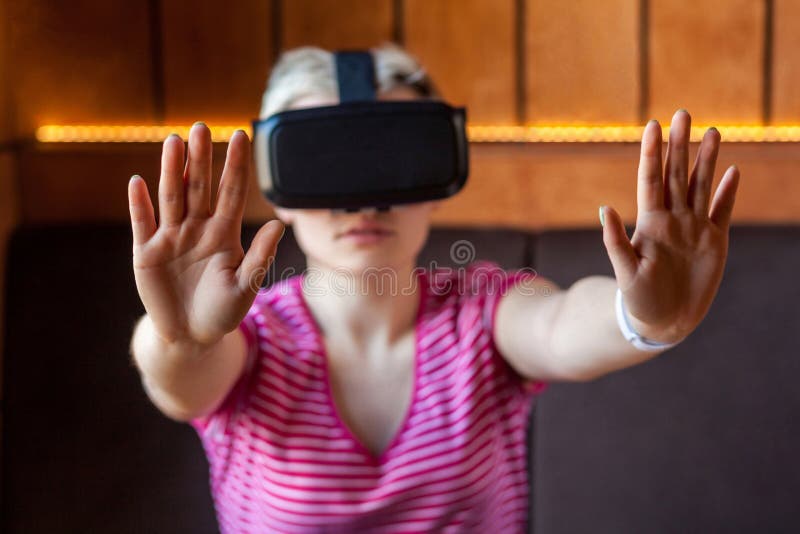 Portrait d'une jeune femme adulte sérieuse vêtue d'un t-shirt rose, assise dans un café, portant un casque de réalité virtuelle e photographie stock
