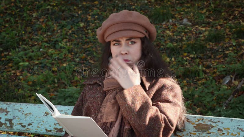 Portrait d'une jeune femme caucasienne en chapeau et manteau lisant un livre dans un parc et montrant des émotions soudaines de dé banque de vidéos