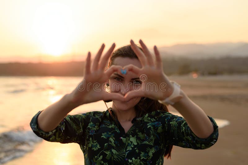 Portrait d'une jeune femme sur la plage au coucher du soleil rouge, coeur des doigts, message de l'amour photo stock Portrait d'une jeune femme sur la plage au coucher du soleil rouge, coeur des doigts, message de l'amour photo stock