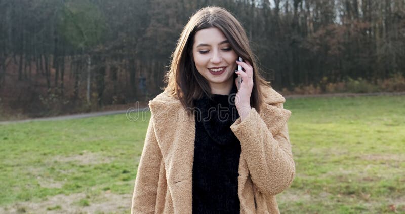 Portrait d'une jeune femme séduisante souriant et décrochant le téléphone et communiquant sur la pelouse verte au parc d'automne banque de vidéos
