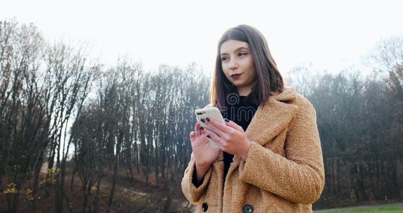Portrait d'une jeune femme séduisante souriant et regardant le smartphone sur la pelouse verte du parc d'automne près de la forêt banque de vidéos