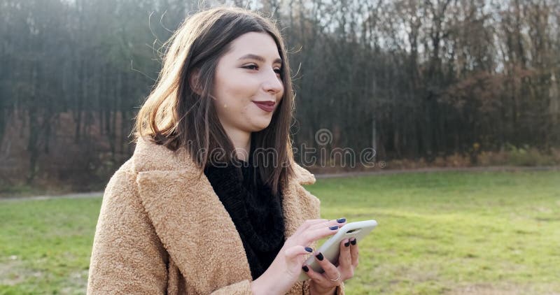 Portrait d'une jeune femme séduisante souriant et regardant le smartphone sur la pelouse verte du parc d'automne près de la forêt banque de vidéos