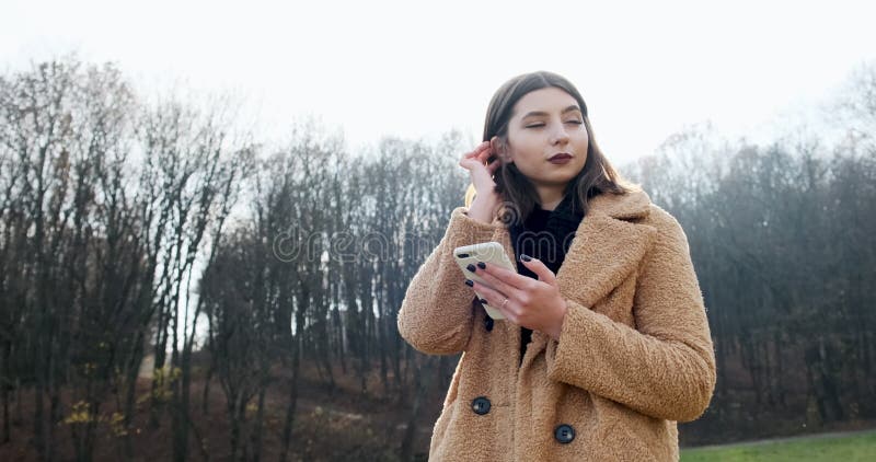 Portrait d'une jeune femme séduisante souriant et regardant le smartphone sur la pelouse verte du parc d'automne près de la forêt banque de vidéos