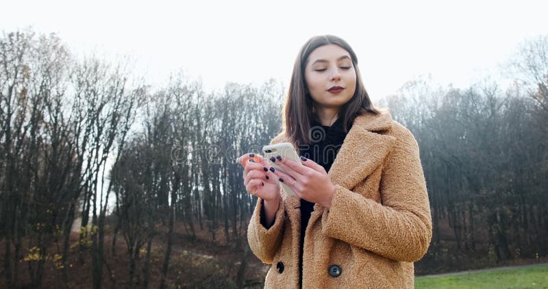 Portrait d'une jeune femme séduisante souriant et regardant le smartphone sur la pelouse verte du parc d'automne près de la forêt banque de vidéos