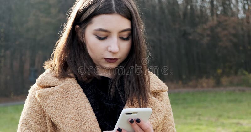 Portrait d'une jeune femme séduisante souriant et regardant le smartphone sur la pelouse verte du parc d'automne près de la forêt banque de vidéos