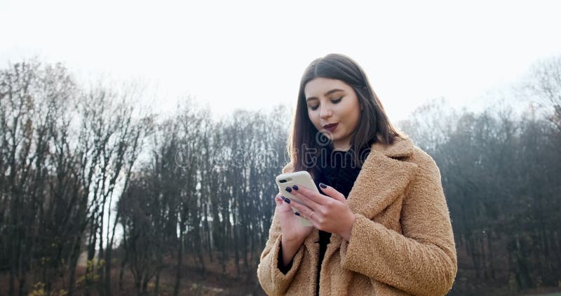 Portrait d'une jeune femme séduisante souriant et regardant le smartphone sur la pelouse verte du parc d'automne près de la forêt clips vidéos