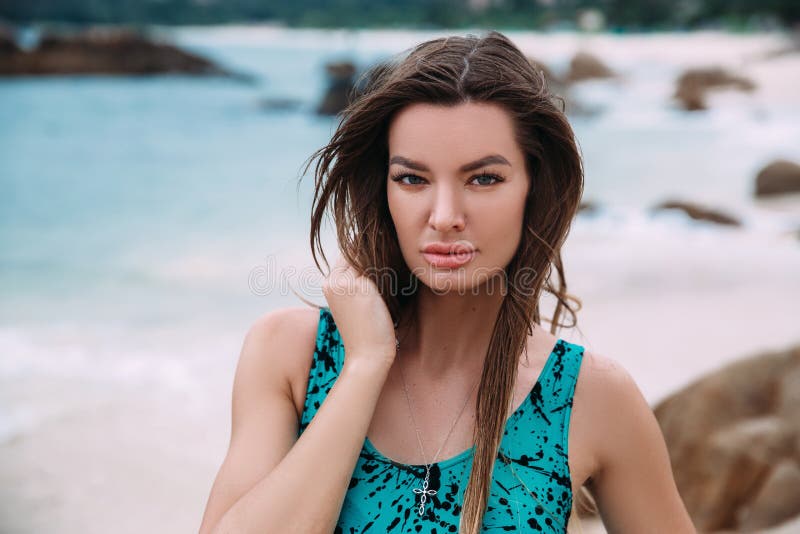 A portrait of a dark-haired, suntanned girl in a bright swimsuit, resting on the azure shore, has an expressive face