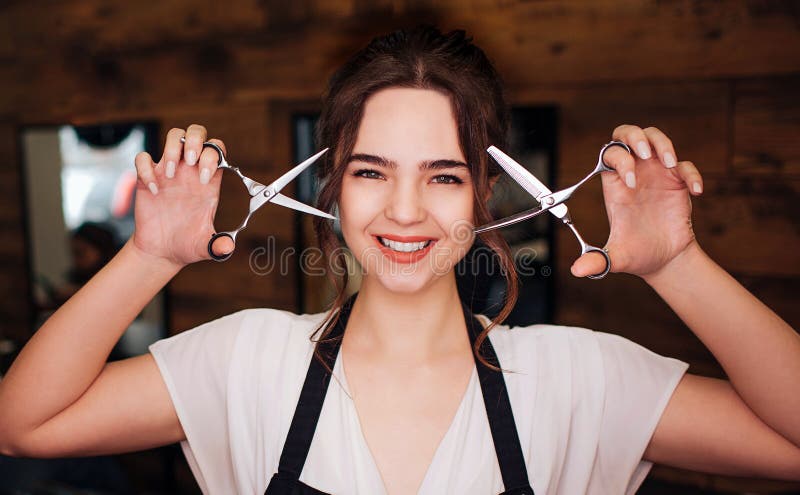 Portrait de belle coiffeuse de sourire de femme avec le tablier noir regardant la caméra tout en tenant des ciseaux professionnel photo libre de droits Portrait de belle coiffeuse de sourire de femme avec le tablier noir regardant la caméra tout en tenant des ciseaux professionnel photo libre de droits