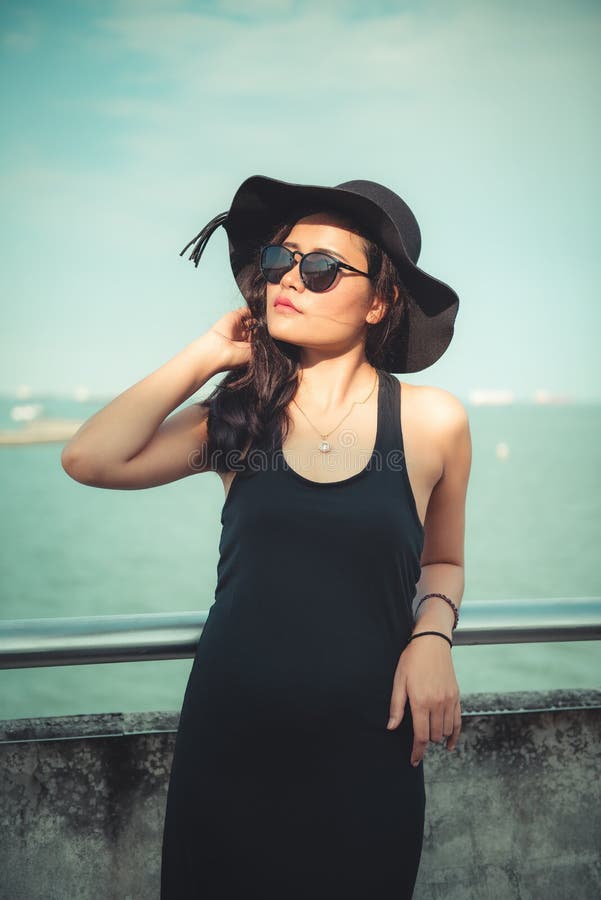 Portrait de belle femme asiatique dans la robe noire de mode avec son chapeau sur le fond de bord de mer, la beauté et le concept photographie stock libre de droits Portrait de belle femme asiatique dans la robe noire de mode avec son chapeau sur le fond de bord de mer, la beauté et le concept photographie stock libre de droits