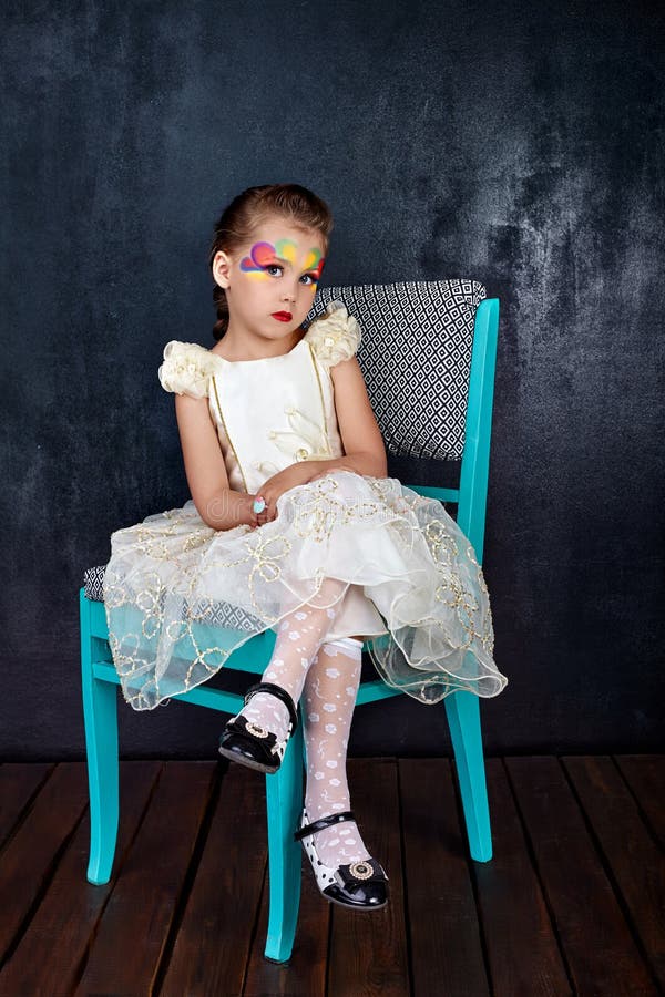 Portrait de belle petite fille dans des lèvres rouges de robe blanche avec le visage peint se reposant sur une chaise au fond fon photos libres de droits Portrait de belle petite fille dans des lèvres rouges de robe blanche avec le visage peint se reposant sur une chaise au fond fon photos libres de droits