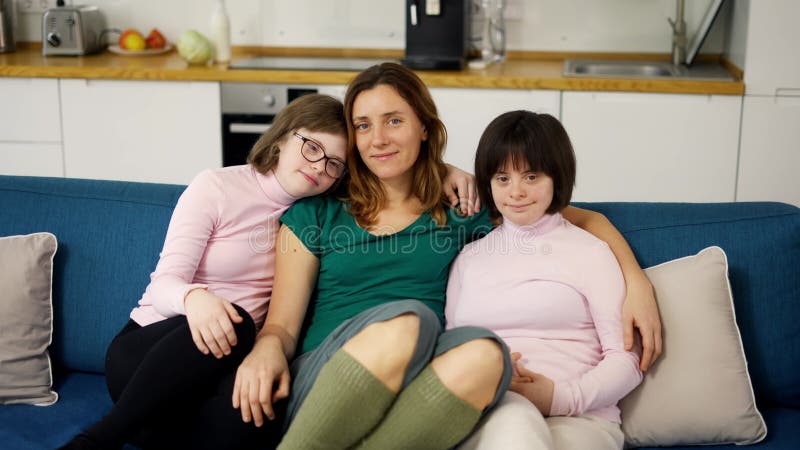 Portrait de deux filles atteintes du syndrome de down assis sur un canapé ensemble embrassant leur mère banque de vidéos
