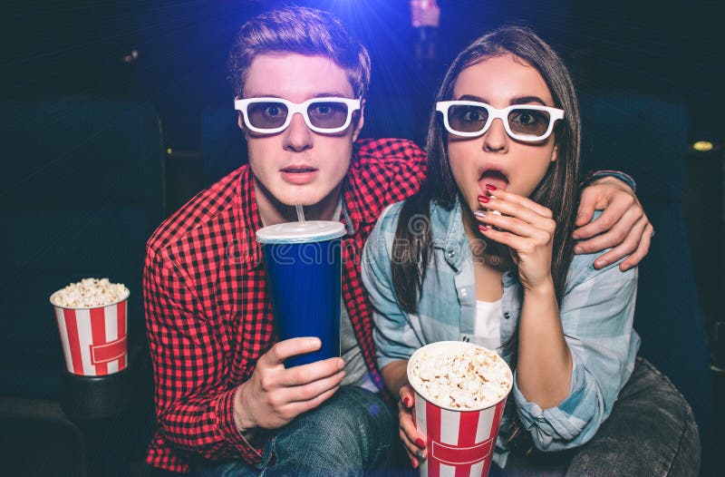 Portrait of two people sitting together in cinema hall and wearing glasses. Girl is amazed and eating popcorn while her partner is drinking cola. Portrait of two people sitting together in cinema hall and wearing glasses. Girl is amazed and eating popcorn while her partner is drinking cola