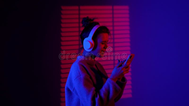 Portrait de femme silhouette dans casque écoutant de la musique sur smartphone stands dans le projecteur isolé sur le néon bleu ro banque de vidéos