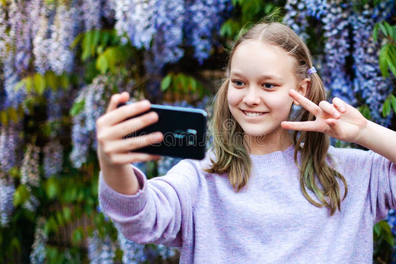 Portrait de fille du caucase de la préadolescence mignonne avec une queue de cheval en tenant le selfie contre la glycine à fleurs photo stock