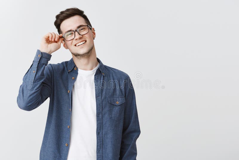 Portrait of nice modern and handsome male student in glasses and blue shirt touching frame of eyewear smiling friendly and delighted, satisfied with productive team work on project over gray wall. Portrait of nice modern and handsome male student in glasses and blue shirt touching frame of eyewear smiling friendly and delighted, satisfied with productive team work on project over gray wall.