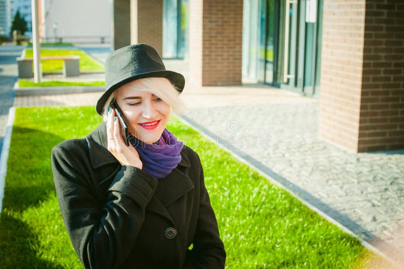 Portrait de jeune belle femme avec les cheveux blancs, dans un manteau noir, une jupe et un chapeau noir, parlant au téléphone po image stock Portrait de jeune belle femme avec les cheveux blancs, dans un manteau noir, une jupe et un chapeau noir, parlant au téléphone po image stock