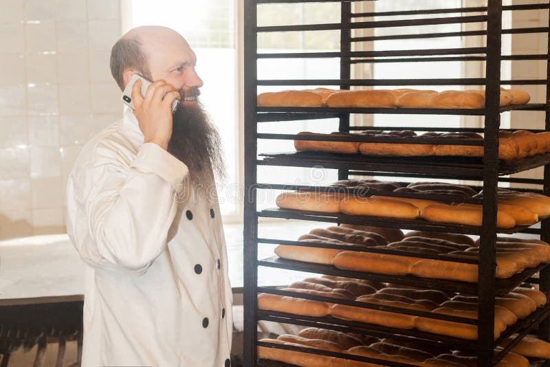 Portrait of happy young adult businessman baker with long beard in white uniform standing in bakery and have online order by phone, talking on cellphone with toothy smile Indoor, profession concept. Portrait of happy young adult businessman baker with long beard in white uniform standing in bakery and have online order by phone, talking on cellphone with toothy smile Indoor, profession concept