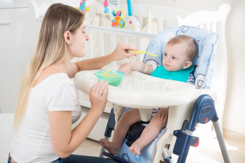 Portrait de jeune mère alimentant son fils de bébé s'asseyant dans le highchair images libres de droits Portrait de jeune mère alimentant son fils de bébé s'asseyant dans le highchair images libres de droits