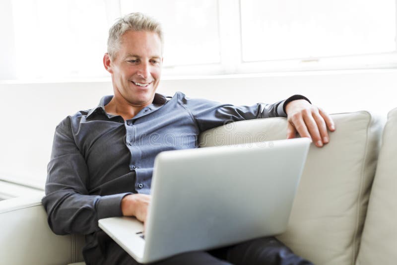 Portrait de l'homme mûr heureux à l'aide de l'ordinateur portable se trouvant sur le sofa dans la maison image stock Portrait de l'homme mûr heureux à l'aide de l'ordinateur portable se trouvant sur le sofa dans la maison image stock