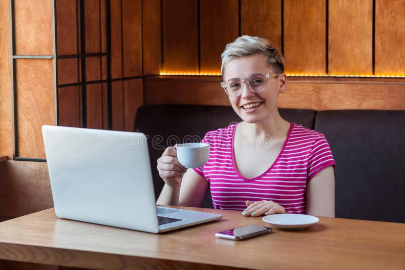 Portrait de la belle jeune femme d'affaires satisfaite aux cheveux courts blonds en t-shirt rose est assise dans un café et boire photographie stock libre de droits