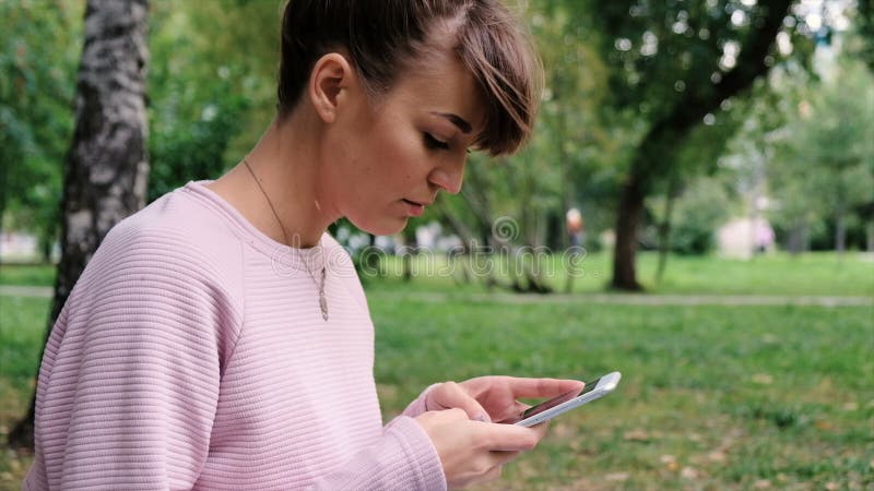 Portrait de la jeune dame décontractée en parc d'été lisant un message textuel à son téléphone portable Belle jeune fille dans le clips vidéos