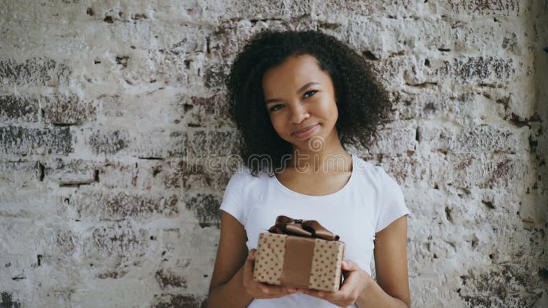 Portrait de la jeune fille africaine heureuse tenant le boîte-cadeau et souriant dans l'appareil-photo clips vidéos