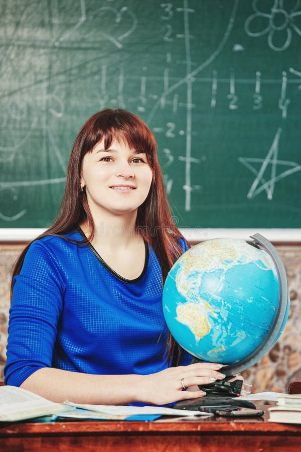 Portrait de professeur la belle jeune femme dans une robe bleue s'assied à un bureau avec un globe sur le fond d'un conseil images stock Portrait de professeur la belle jeune femme dans une robe bleue s'assied à un bureau avec un globe sur le fond d'un conseil images stock